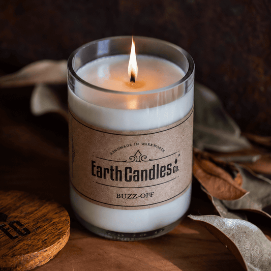 Lit Buzz-Off soy candle by Earth Candles Co. in a cut and polished upcycled wine bottle, with dried leaves and the wooden lid alongside.