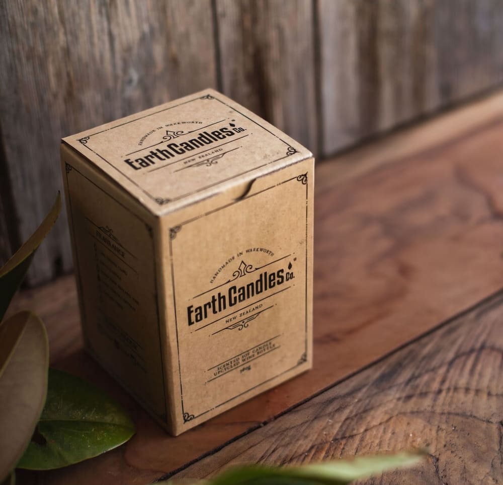 The kraft paper retail box that Earth Candles Co. wine bottle soy candles are packed in, on a rustic wooden surface with green leaves in the foreground.