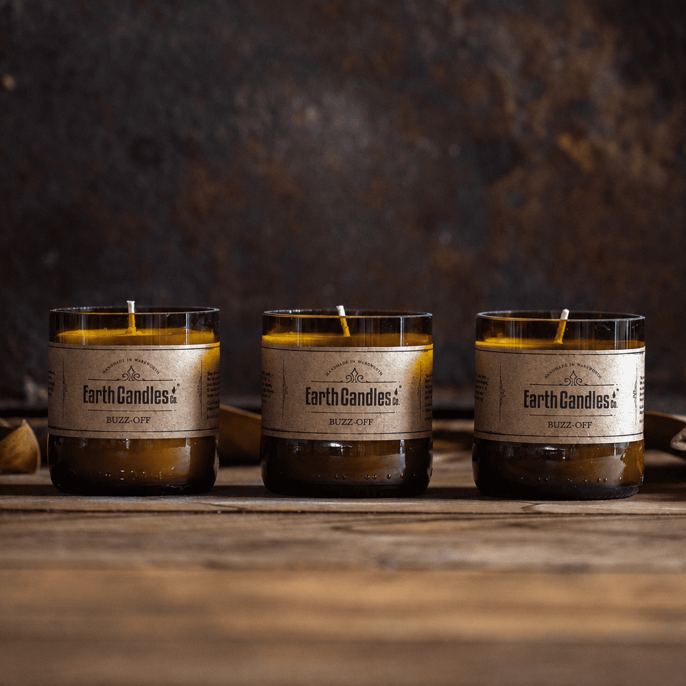 Three Buzz-Off soy tealights by Earth Candles Co., unlit, in cut amber beer bottle bases on a wooden bench