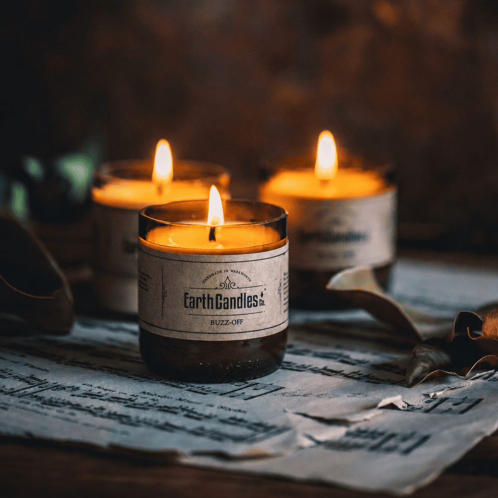 Three Buzz-Off soy tealights burning in cut amber beer bottle bases, on sheet music with dried leaves