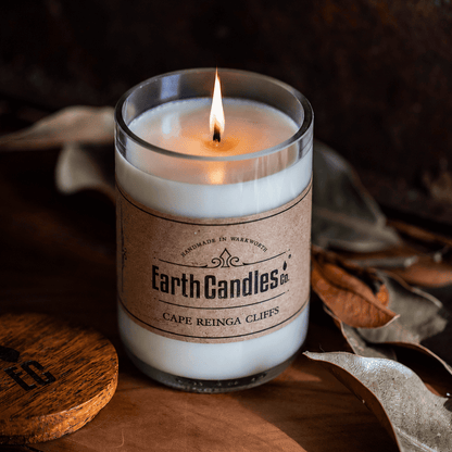 Lit Cape Reinga Cliffs soy candle by Earth Candles Co. in a cut and polished upcycled wine bottle, with dried leaves and the wooden lid alongside.