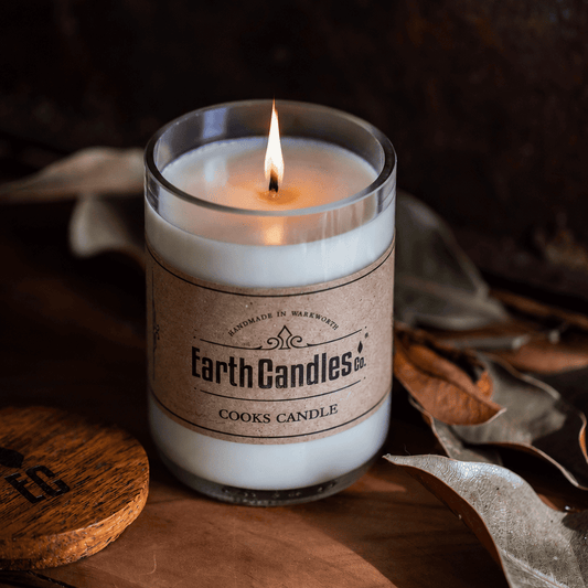 A lit Earth Candles Co. Cooks Candle soy candle in an upcycled wine bottle, cut and polished, with a kraft paper label, on a rustic wooden surface with dried leaves and the wooden lid beside it.