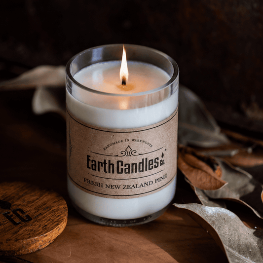 Lit Fresh New Zealand Pine soy candle by Earth Candles Co. in a cut and polished upcycled wine bottle, with dried leaves and the wooden lid alongside.