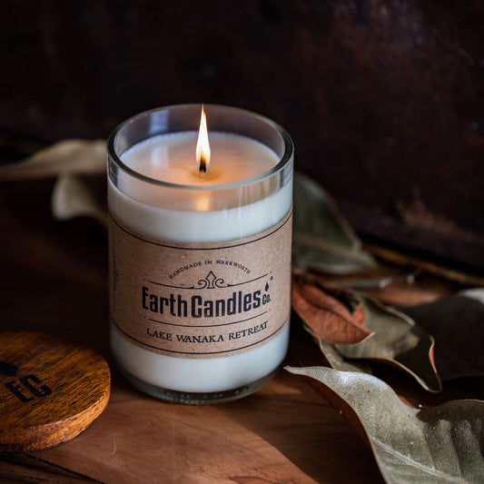 Lit Lake Wanaka Retreat soy candle by Earth Candles Co. in a cut and polished upcycled wine bottle, with dried leaves and the wooden lid alongside.