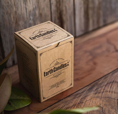 The kraft paper retail box that Earth Candles Co. wine bottle soy candles are packed in, on a rustic wooden surface with green leaves in the foreground.