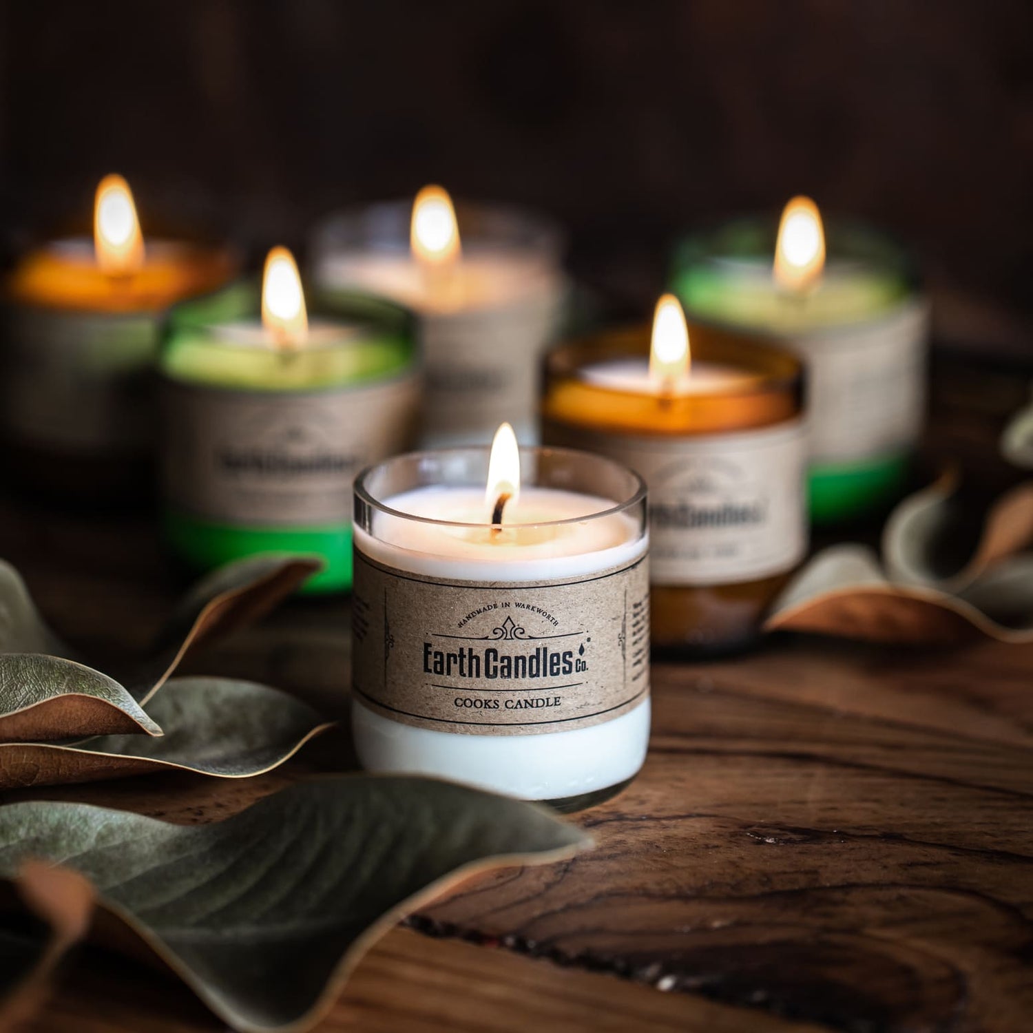 Six lit Earth Candles soy tealights in clear, green and amber beer bottle bases on a dark wooden table
