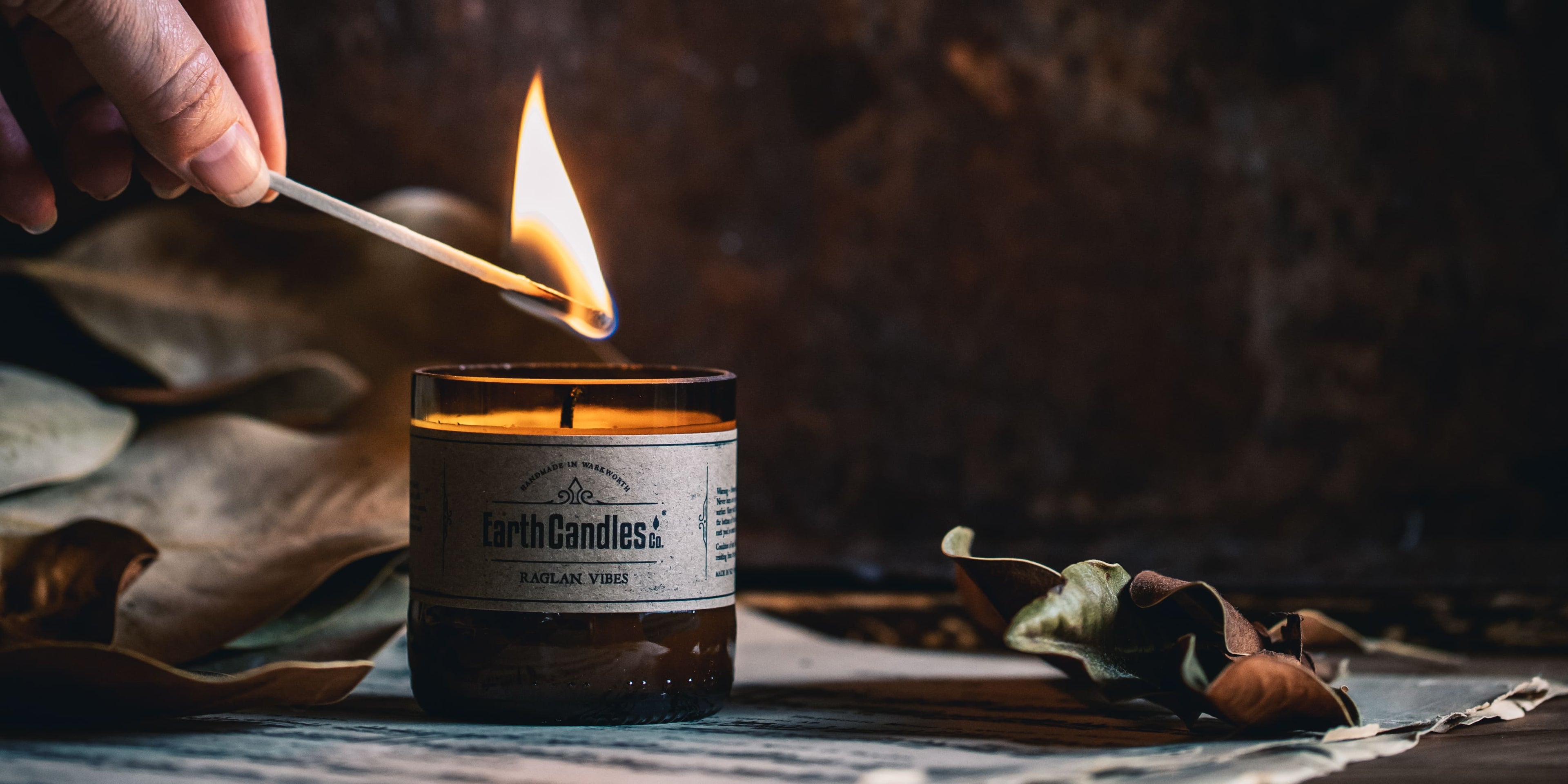 A hand lighting an Earth Candles Co. Raglan Vibes soy candle with a match, in a cut amber bottle with a kraft label, on newspaper with dried leaves.