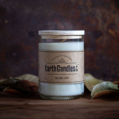 A re-purposed wine bottle soy candle with a label reading 'Earth Candles Buzz-Off', surrounded by leaves on a wooden surface.