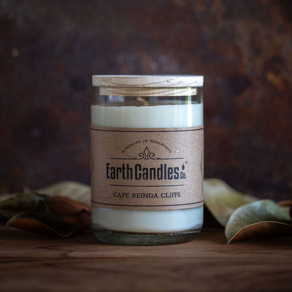A soy wax candle in a clear re-purposed wine bottle with a brown label that reads 'Earth Candles - Cape Reinga Cliffs'. The candle is on a wooden surface with leaves around it.