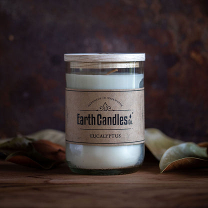 A white soy wax candle in a clear upcycled wine bottle with a brown label that reads 'Earth Candles Eucalyptus'. The candle is surrounded by eucalyptus leaves on a wooden surface.